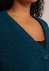 Teal fluffig cardigan med V-ringning och enkel knappstängning, med en mjuk textur och synliga sömmar längs kanterna.