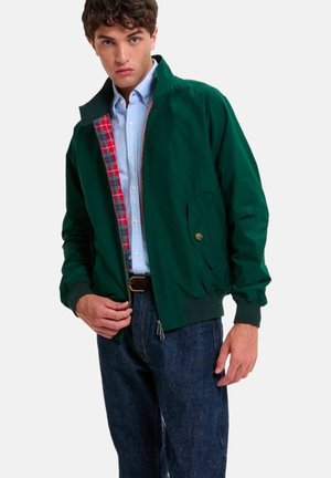 Giovane uomo che indossa una giacca verde scuro con zip e interno a quadri rossi, camicia azzurra, jeans scuri e cintura marrone, in piedi su sfondo bianco.