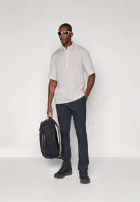 Polo a maniche corte grigio chiaro con tasca, abbinata a pantaloni blu navy e zaino nero. Scarpe nere di rilievo e occhiali da sole con montatura dorata.