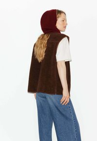 Gilet en polaire marron avec des côtés ouverts, porté sur un t-shirt blanc. Associé à un jean en denim ample et à une capuche rouge en tricot. Design simple.