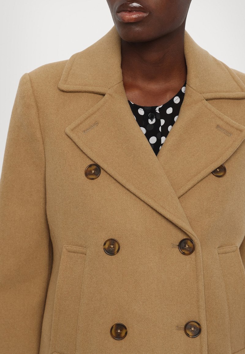 Beige doppelt-knöpfiger Wollmantel mit großen braunen Knöpfen, Reverskragen und einer vertikalen Tasche, über einer schwarzen Bluse mit Punkten getragen.