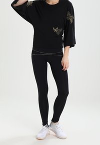 Czarny, oversize top z złotym haftem motyla, zestawiony z czarnymi legginsami i białymi butami sportowymi. Gładki materiał, luźny krój, codzienny design.