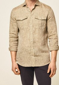 Chemise à manches longues marron clair avec deux poches poitrine, fermeture boutonnée, et poignets retroussés. Confectionnée en tissu texturé.