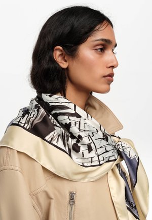 Joven con chaqueta beige y una bufanda de seda estampada en negro, blanco y crema atada alrededor del cuello, vista de perfil hacia la derecha.