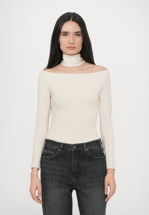 Off-white long-sleeve top with a mock neck, made from smooth material; paired with high-waisted dark gray jeans featuring front pockets.