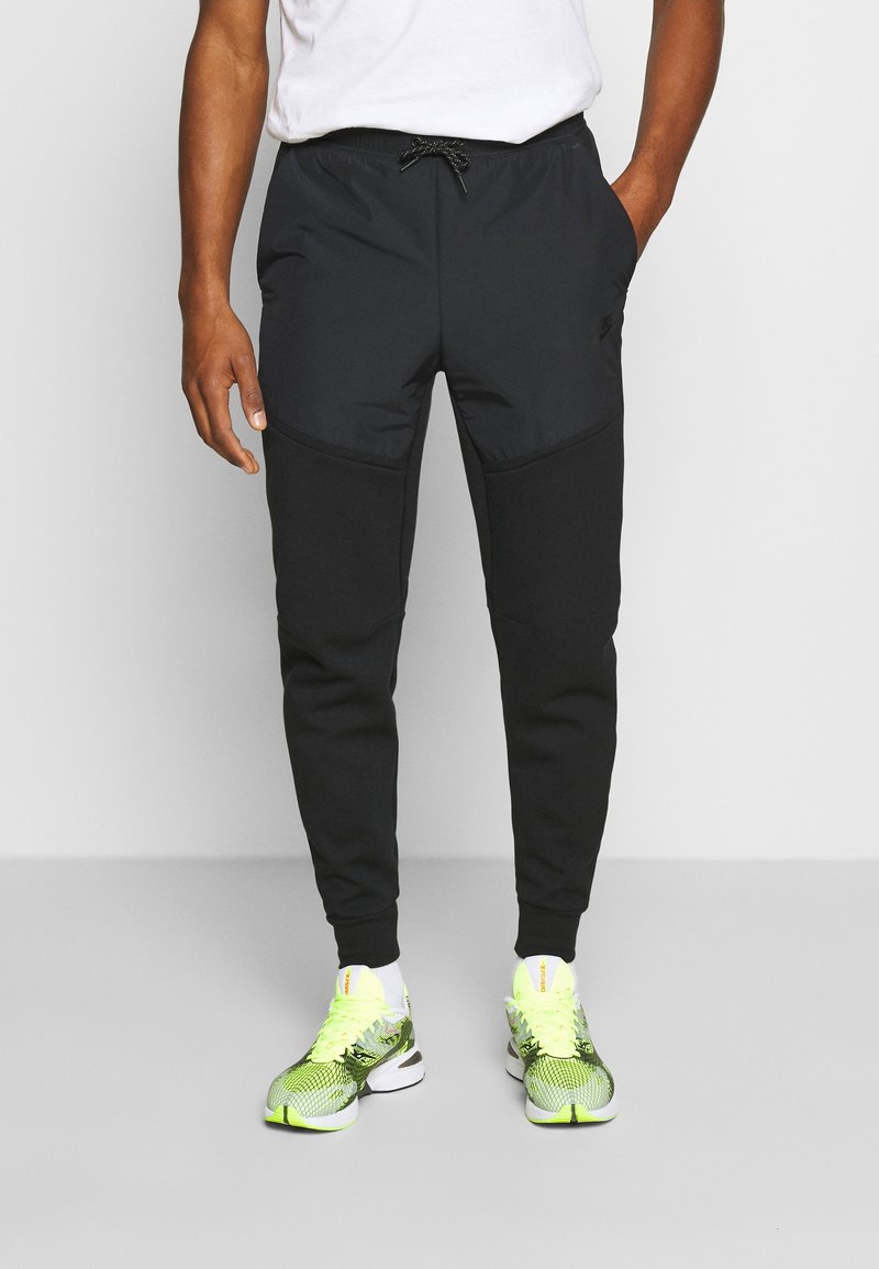 Černé sportovní tepláky z hladké, pružné látky, s páskem na stažení v pase, zúženým střihem a kontrastními texturami v oblasti kolen.