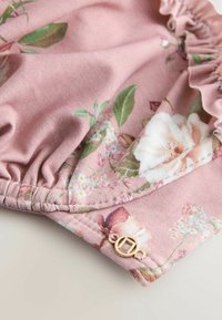 Tissu floral rose avec bordure à volants et petit charm circulaire doré sur une surface claire.