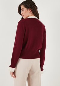 Bordový svetr s žebrovaným lemem a manžetami, s krajkovými detaily na manžetách. V kombinaci se světle béžovými kalhotami. Hladká textura.