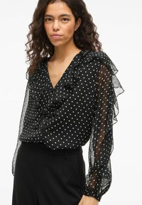 Blusa nera con pois bianchi, con dettagli arricciati, maniche lunghe trasparenti e scollatura a V. Presenta dettagli con bottoni e una vestibilità comoda.