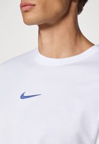 Hvid bomulds t-shirt med et blåt broderet Nike-logo på venstre bryst. Rund halsudskæring og glat stofstruktur.
