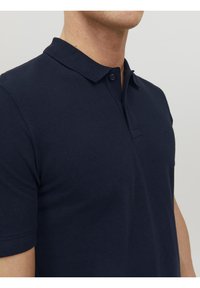 Homme portant un polo bleu marine à manches courtes avec un col plié et une patte à deux boutons, montré des épaules au cou.