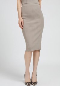 Frau in einem beigen, gerippten Bleistiftrock und spitzen, leopardgemusterte High Heels steht vor einem schlichten Hintergrund.