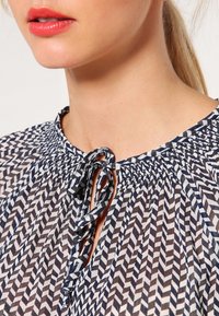 Blusa leggera navy e bianca con stampa a motivo, colletto arricciato e dettaglio con fiocco; presenta una stampa geometrica e una texture trasparente.