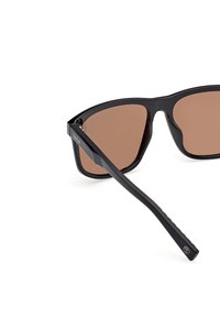 Schwarze Sonnenbrille mit einem schlanken, eckigen Rahmen, braun getönten Gläsern und strukturierten schwarzen Bügeln mit einem Logo. Ideal für UV-Schutz.