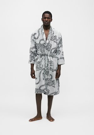 Άνδρας που στέκεται ξυπόλυτος φορώντας μια γκρι ρόμπα μπάνιου με ζώνη και μαύρα μοτίβα λεοπάρδαλης και ουράς σε λευκό φόντο.