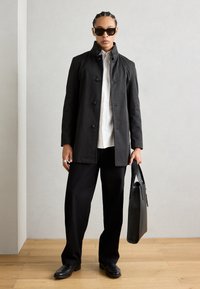 Zwarte waterdichte jas met knoopsluitingen, gedragen over een wit shirt. In combinatie met losse zwarte broeken en een rechthoekige zwarte tas.