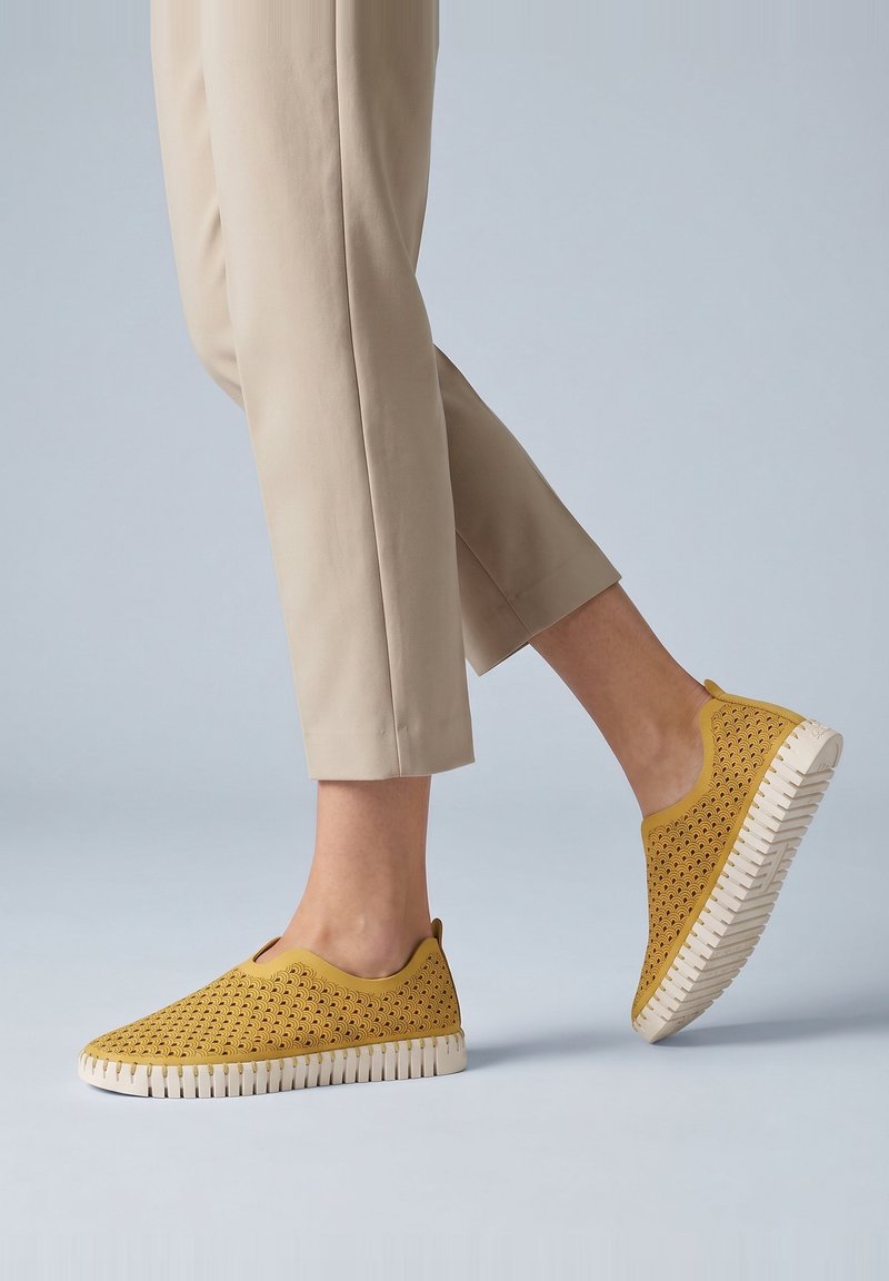 Piernas con pantalones beige y zapatos tipo mocasín amarillos mostaza con suelas blancas gruesas y ranuradas pisando un suelo gris claro.