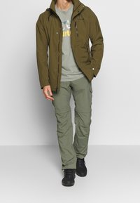 Olivgrön vattentät jacka med justerbar huva, ficka med dragkedja på bröstet, tillsammans med ljusgröna cargobyxor och svarta skor.