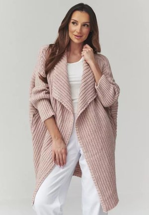 Kobieta ubrana w długi, gruby różowy kardigan z dzianiny, na białej bluzce i białych spodniach, stojąca na jednolitym jasnym tle.