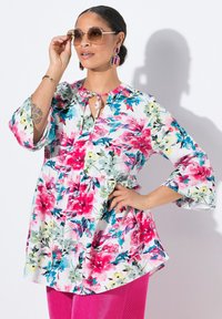 Blumenbluse aus leichtem Stoff mit lebhaften pinken, blauen und grünen Mustern. Mit einem Schlüssel-Ausschnitt und ausgestellten Ärmeln, kombiniert mit pinken Cordhosen.