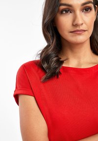 Haut rouge à manches courtes avec une texture lisse et des manches retroussées, présentant un col rond classique et une coupe décontractée.