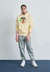 Sweatshirt amarelo da Nike com gráfico, calças de fato cinzentas e ténis brancos. O hoodie tem um bolso frontal e punhos canelados.
