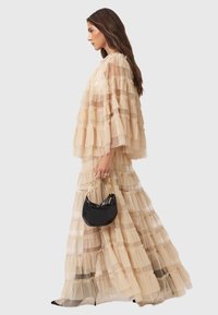 Μαξι φόρεμα από τulle σε νουντ, με ριγέ tiers και διάφανα μανίκια. Συμπληρωμένο με μια μαύρη τσάντα χειρός με χρυσά μεταλλικά στοιχεία.