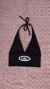 Μαύρο τοπ με τιράντες που δένει στον λαιμό, κατασκευασμένο από ελαστικό ύφασμα. Διαθέτει βαθύ V-λαιμόκοψη και κεντημένο λευκό λογότυπο στο εμπρόσθιο μέρος.