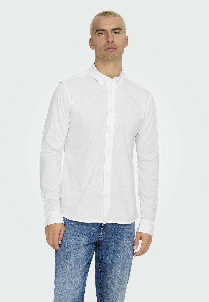 White long-sleeve shirt with button-down collar, made of smooth fabric, paired with blue jeans, model stands against a light background.