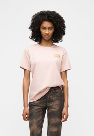 Mladá žena s kučeravými vlasmi v svetloružovom tričku Diesel a vyblednutých čiernych džínsoch stojaca pred jednoduchým pozadím.