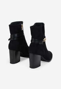 Botines de ante negro con un alto tacón en bloque, cremallera lateral y una correa de cuero negro con un detalle de hebilla dorada.