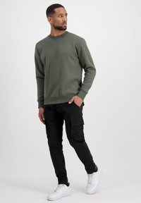 Felpa verde oliva con scollo a girocollo, abbinata a pantaloni cargo neri con tasche e sneakers bianche. Texture del tessuto liscia.