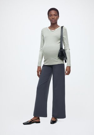 Těhotná žena v proužkovaném tričku s dlouhým rukávem, širokých tmavých kalhotách, černých balerínách a černé kabelce přes rameno na jednobarevném bílém pozadí.