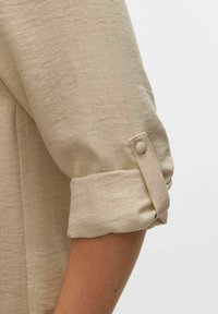 Beige linnen shirt met opgerolde mouwen, voorzien van een knoopband voor een veilige aanpassing van de manchetten. Soepele textuur met subtiele weefdetails.