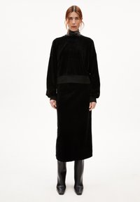 Schwarzes Samtkleid mit hohem Kragen und langen Ärmeln, mit taillierter Taille und gerader Silhouette, getragen mit schwarzen knielangen Stiefeln.