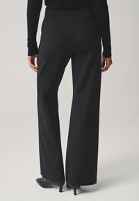 Svarta vida byxor i glatt tyg med dold midjeresår, försedd med bakfickor och kombinerade med svarta högklackade skor.