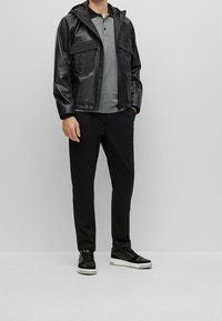 Svart läderjacka med huva, grå kortärmad polo, svarta byxor och svarta sneakers med vita sulor, mot en ljus bakgrund.