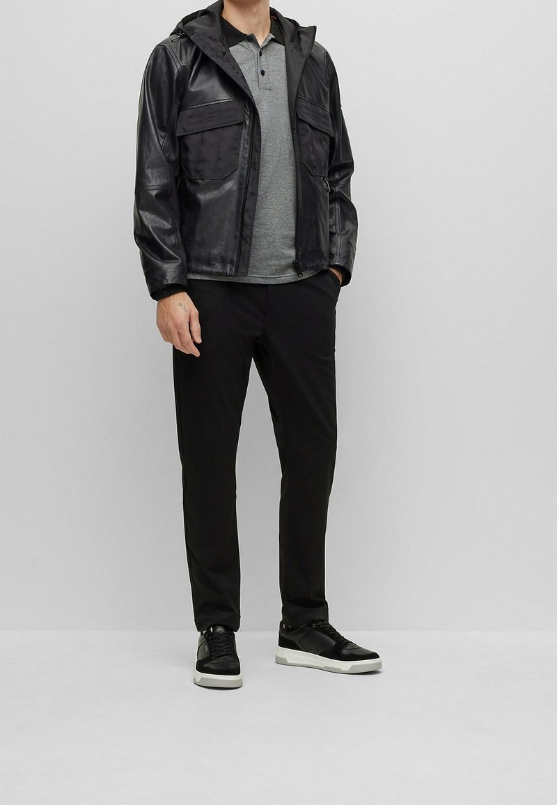 Svart läderjacka med huva, grå kortärmad polo, svarta byxor och svarta sneakers med vita sulor, mot en ljus bakgrund.