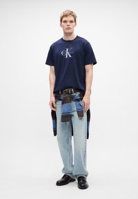 Námornická polokošile s krátkým rukávem s bílým logem "Calvin Klein", světle modré džíny, hnědý pásek a modro pruhovaný svetr uvázaný v pase. Černé boty.
