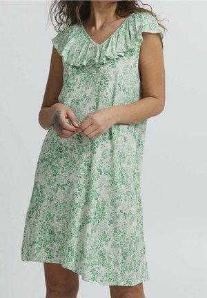 Femme portant une robe blanche à manches courtes, longueur genoux, avec un motif floral vert et un col volanté, debout les mains jointes.