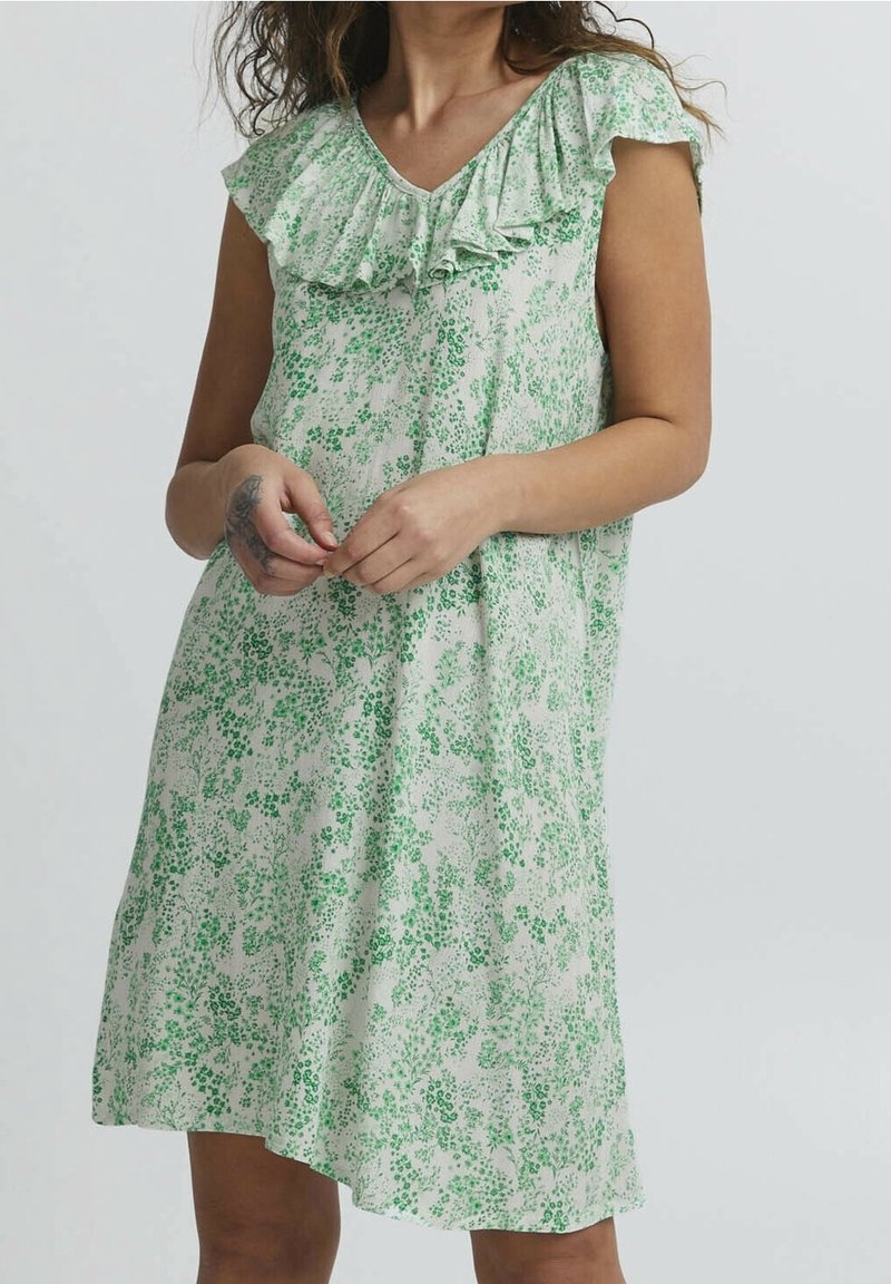 Femme portant une robe blanche à manches courtes, longueur genoux, avec un motif floral vert et un col volanté, debout les mains jointes.