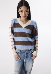 Pull cropped rayé en bleu, marron et crème. Avec un col en V et une texture lâche. Associé à un jean bleu déchiré.