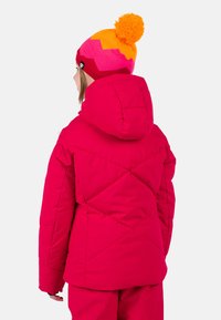 Vaaleanpunainen puffer-takki hupulla, jossa on timanttikudottu kuvio ja joustavat hihansuut. Yhdistetty moniväriseen neulottuun lämpöpäähineeseen, jossa on pom-pom.