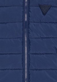 Vest puffer albastru marin cu panouri cusute orizontal, închidere centrală cu fermoar și un patch texturat negru cu logo pe piept.