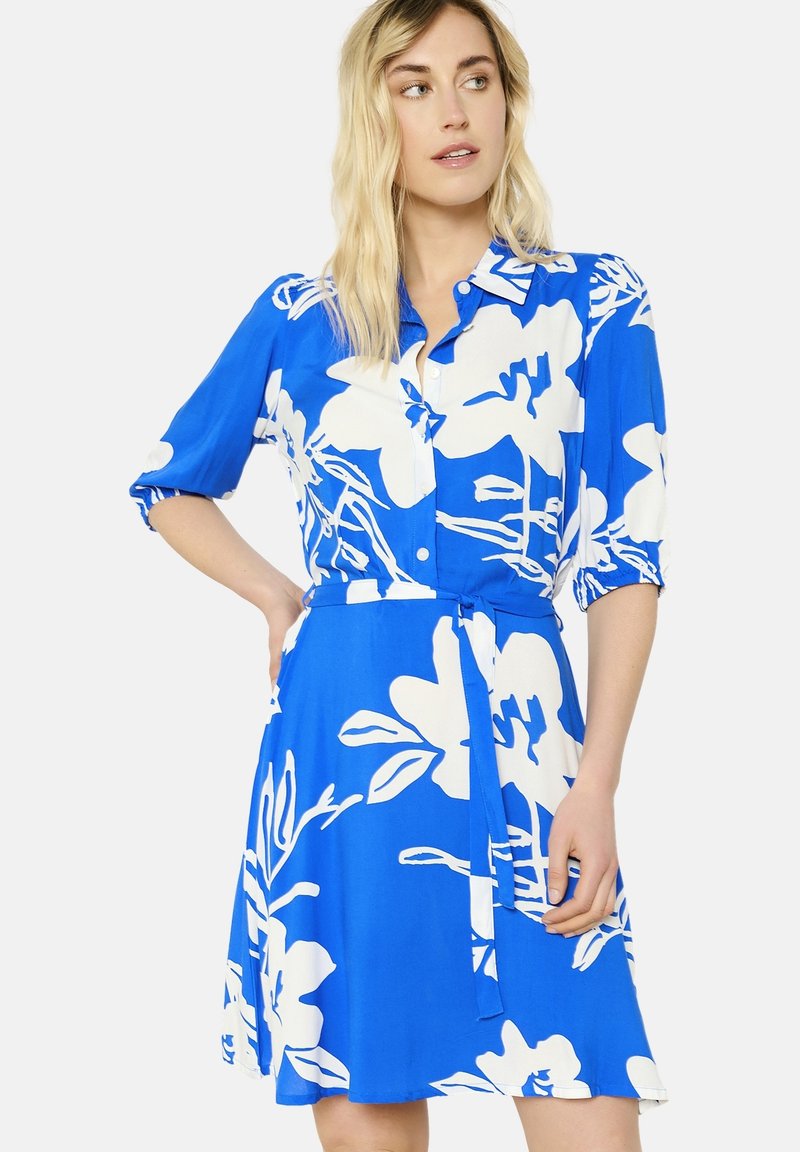 Sininen mekko, jossa on valkoinen kukkakuosi, napitus edessä, lyhyet hihat ja vyö vyötäröllä. Sileä kangas ja polven pituinen muotoilu.