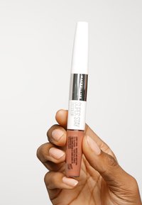 Płynna szminka Maybelline Super Stay w przezroczystym opakowaniu w różowo-brązowym odcieniu, z białą nakrętką z srebrnymi akcentami.