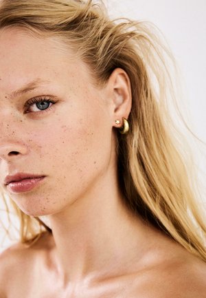 Približeni prikaz svijetlopute žene s plavim očima, pjegicama, plavom kosom i dvije zlatne naušnice u obliku zvijezde i polumjeseca.