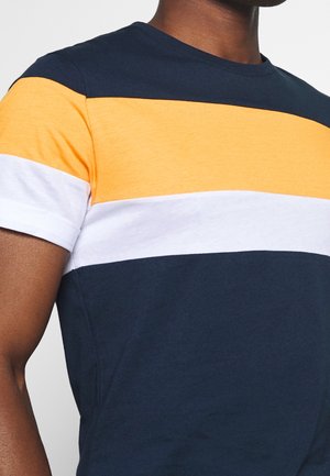 Mann trägt ein T-Shirt mit kurzen Ärmeln und horizontalen Streifen in Marineblau, Gelb und Weiß, gezeigt vom Brustbereich aufwärts.