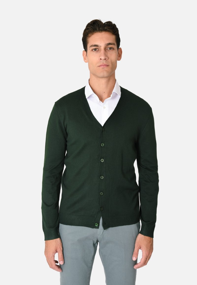 Cardigan verde scuro a V in tessuto morbido, con chiusura a bottoni, polsini a coste e orlo dritto. Indossato sopra una camicia bianca.