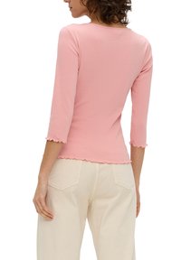 Roze aansluitend topje met driekwartrmouwen en een afgeronde zoom. Gemaakt van zachte, gestructureerde stof. Gecombineerd met lichtbeige broeken.